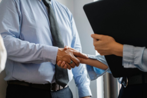 Two professionals shaking hands in an office setting, one holding a folder or tablet.