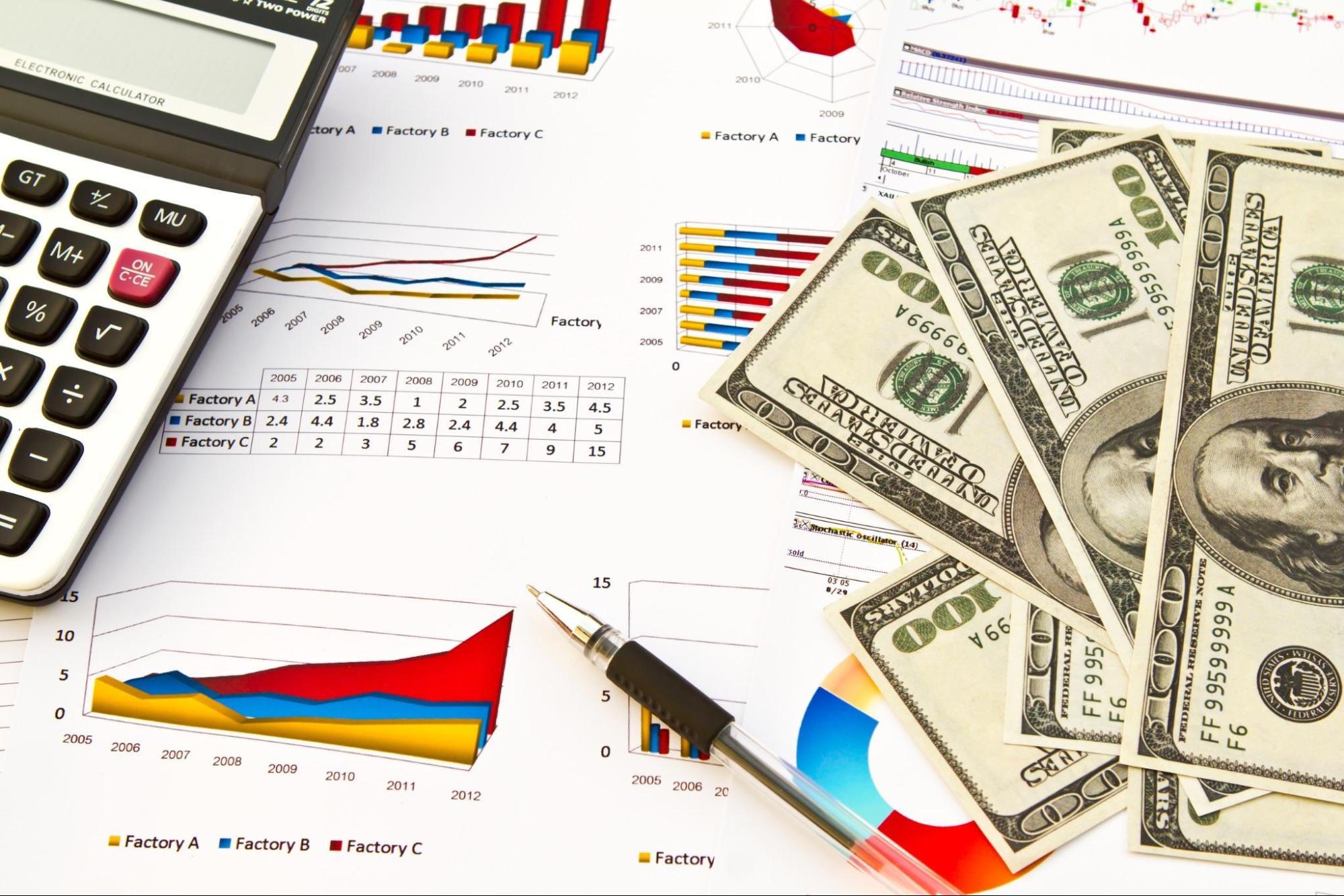 A few hundred dollar notes and a calculator and a pen is kept on several sheets containing data tables, diagrams and charts. Caption: Spend management helps SMBs reduce unnecessary business expenses