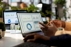 A business analyst pointing at a financial dashboard on a laptop displaying charts, donut graphs, and liquidity metrics, with a second monitor showing bar charts in the background.